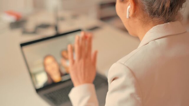Businesswoman in white blazer smiles and waves during group video call on laptop with diverse colleagues discussing work.