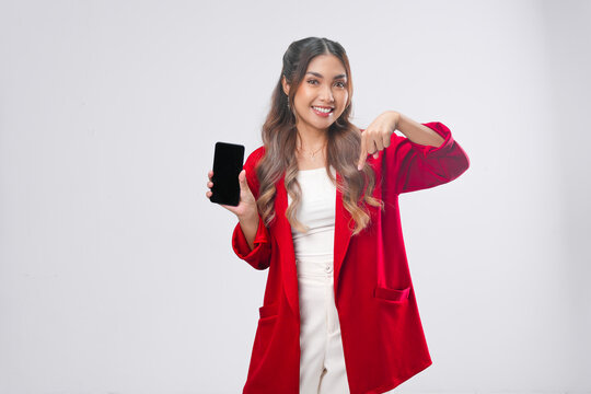 Joyful young Asian woman in red jacket holding smartphone while pointing to the copy space below.