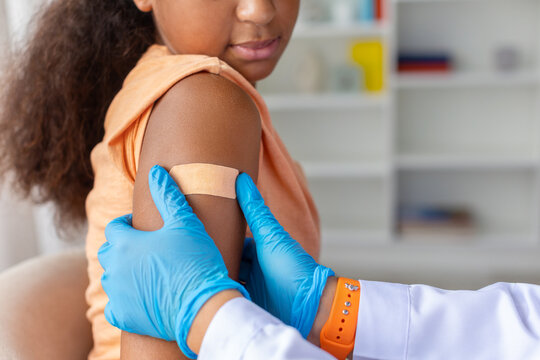 Closeup of young Black girls upper arm with adhesive bandage after vaccination as medical professional in gloves provides careful post-injection aftercare