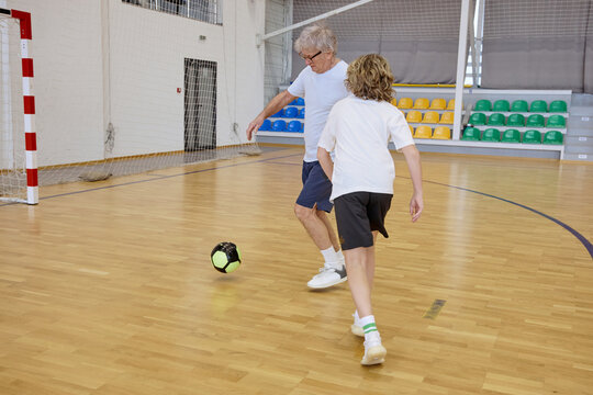 Senior man and teenage boy kicking ball during an active session in an indoor sports hall