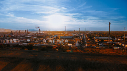 Fototapeta premium Industrial site during sunset with facilities and structures present. A large industrial area shows many buildings and pipes under a sunset sky.