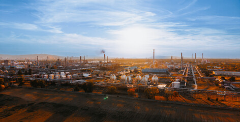 Fototapeta premium Sunset view over petroleum refinery and gas storage spheres. Sun hangs above distant mountains, illuminating central spherical containers, long dark shadow field in foreground.