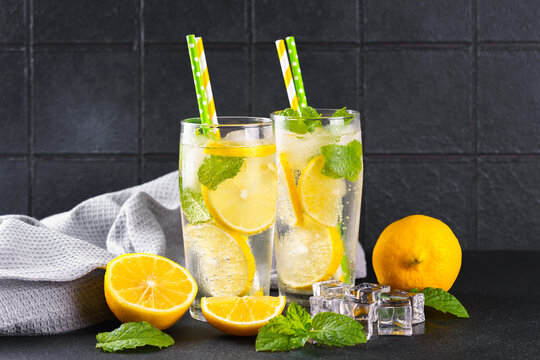 Close up of two clear glasses with refreshing homemade lemonade with fresh lemon slices, mint leaves, ice cubes and paper straws on a light kitchen counter against black tile background, copy space