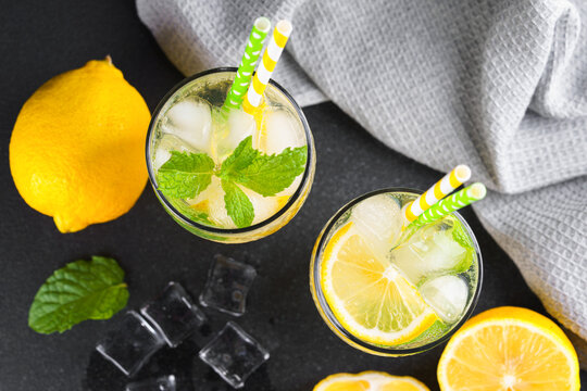 Top view of refreshing homemade lemonade in glasses with fresh mint leaves, lemon slices, ice cubes and soda water and paper straws on a black background. Summer citrus drink, cold icy beverage.