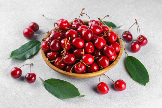 Fresh ripe red sweet cherries in wooden bowl with green leaves on light textured background. Healthy organic summer fruit, juicy red berries, natural freshness, healthy eating, seasonal food