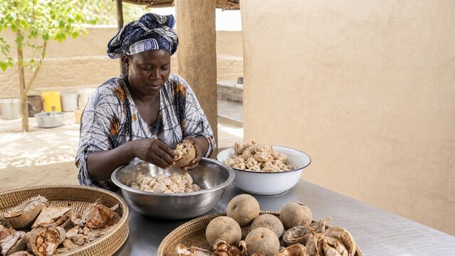 A Nigerian woman market vendor smiling while sitting near fresh produce like yams and grains, showcasing local commerce and rich cultural life, vibrant street scene energy