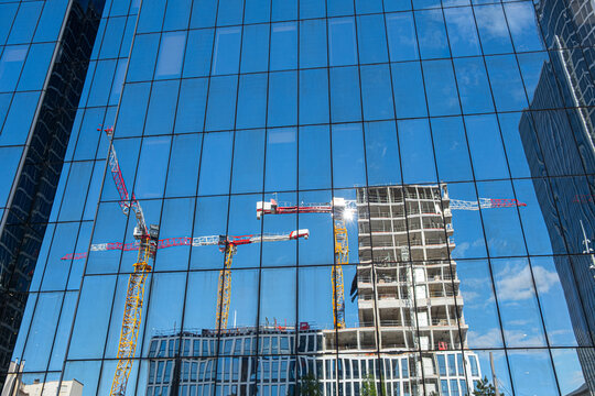 Across a blue facade, reflection layers construction across the view as glass holds framework near center, while cranes frame an unfinished tower and visible development