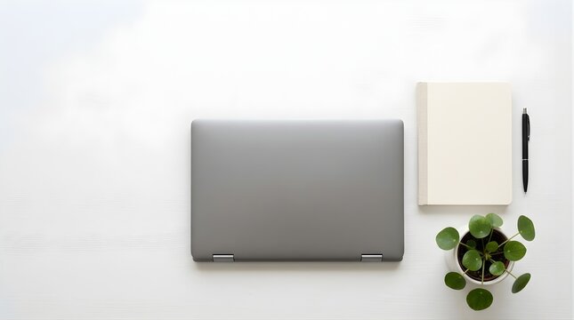 Closed grey laptop with notebook and plant on a white desk top view