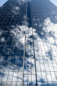 Across a blue facade, shifting clouds appear in reflection, while beyond them a glass grid defines the skyscraper as vertical windows rise toward open sky, emphasizing depth