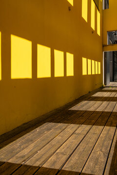 Across the frame, a yellow wall catches sunlight beside the walkway, while warm rectangles from windows stretch as shadows over wooden boards in a measured rhythm
