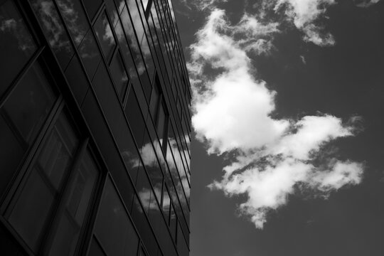 Looking upward, a monochrome skyscraper facade rises through the frame, vertical lines in glass catching light before clouds fill the sky and reflection deepens contrast