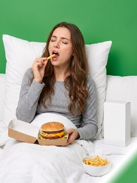 Hungry young woman sitting comfortably in bed while eating delicious golden french fries and holding a large juicy cheeseburger against a vibrant solid green background