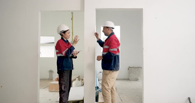 Asian woman engineer and male contractor discuss wall measurement inside unfinished room, reviewing construction safety compliance, building progress, quality control, work instruction indoors.