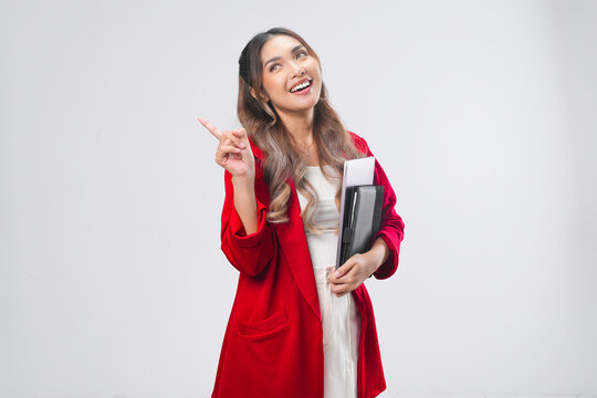 Happy young Asian woman holding laptop and book while pointing to the copy space behind her.