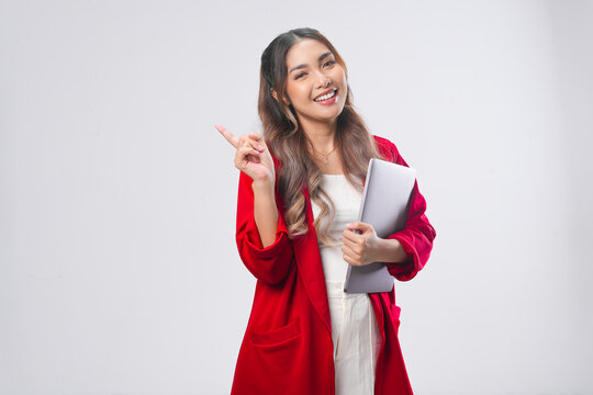 Cheerful young Asian woman holding laptop while pointing to the copy space behind her.