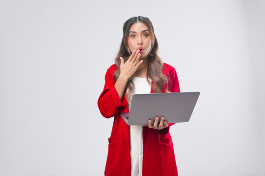 Surprised young Asian woman in a red jacket holding her laptop with her mouth wide open.