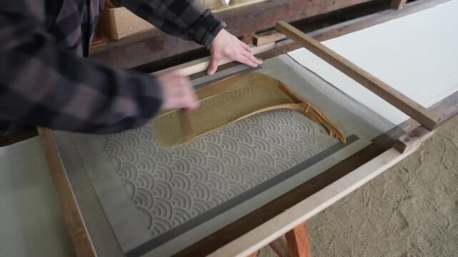 Wave patterned stencil screen lowered onto white cloth as the dyer steadies the timber frame and readies the resist paste