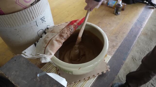 Wooden paddle stirring thick brown rice resist paste inside a large ceramic pot, the mixture falling in glossy ribbons
