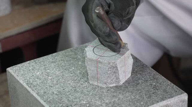 Gloved hands tap a chisel with a mallet to shape a small hexagonal granite blank resting on a polished stone workbench