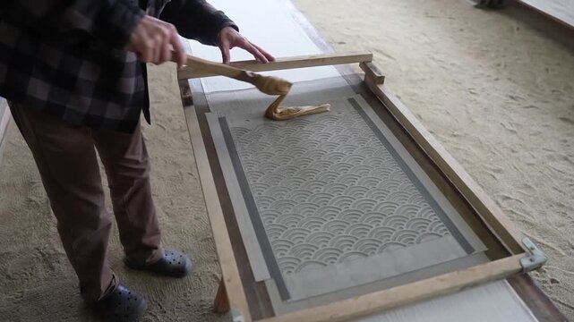 Dyer crosses a sand floored workshop with a wooden spatula and sets a wave patterned stencil frame over a long white cloth