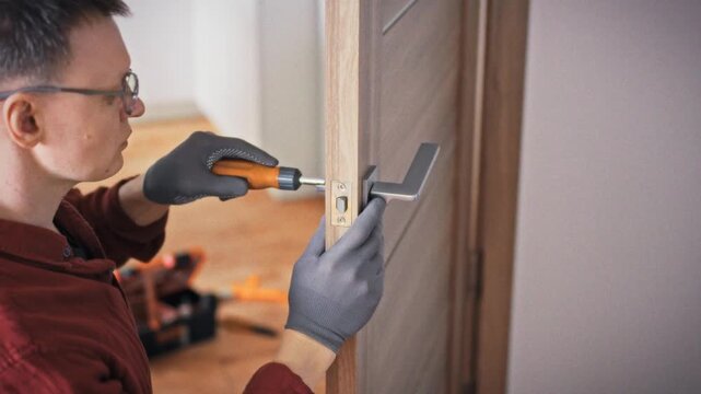 A close-up of a technician's face in glasses focused on repairing a door lock mechanism with an orange screwdriver on a wooden interior door. Concept of skilled and precise home repair work