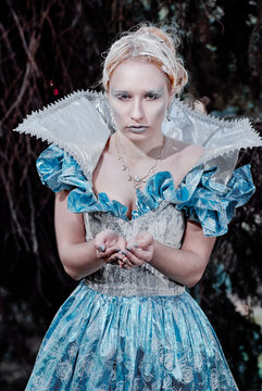 Ethereal ice queen portrait wearing blue vintage gown and glittery frost makeup, cupping powdery snow in her hands against dark woodland backdrop, conveying cold elegance, mystique and regal fantasy.