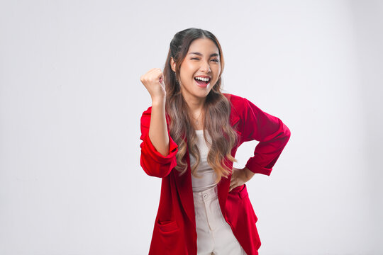 Cheerful young Asian woman in a red jacket is raising her fist with happy successful expression.
