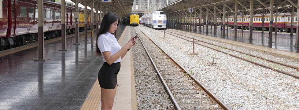 A young Asian woman with long black hair in a white short-sleeved university uniform and black skirt looks at her smartphone