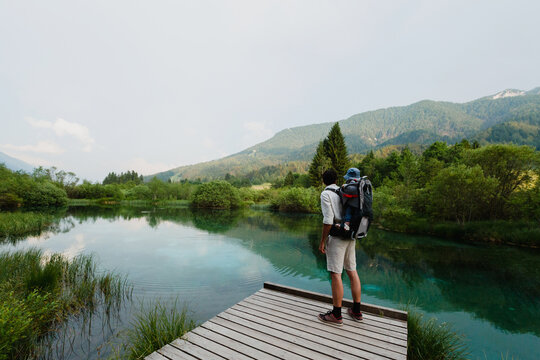 A father carrying his baby in a backpack stands on a wooden pier looking at a pristine alpine lake. Active family travel concept highlighting ecological tourism and exploring natural landscapes.