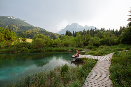 A woman practices outdoor meditation on a wooden boardwalk beside an emerald alpine lake. Harmonious eco-tourism concept focusing on spiritual connection with beautiful natural landscapes.