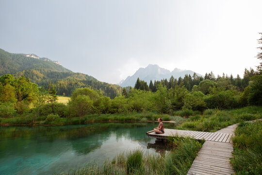 A woman practices outdoor meditation on a wooden boardwalk beside an emerald alpine lake. Harmonious eco-tourism concept focusing on spiritual connection with beautiful natural landscapes.