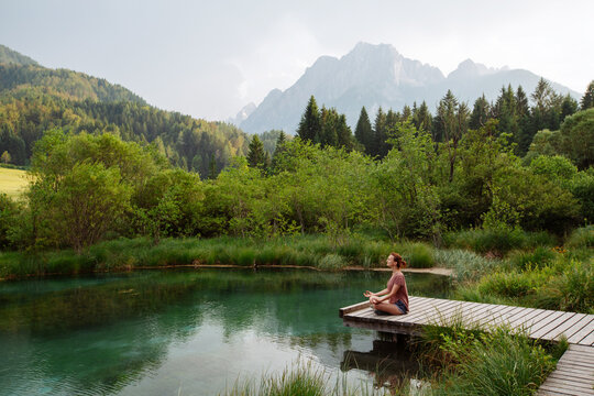 A woman practices outdoor meditation on a wooden boardwalk beside an emerald alpine lake. Harmonious eco-tourism concept focusing on spiritual connection with beautiful natural landscapes.