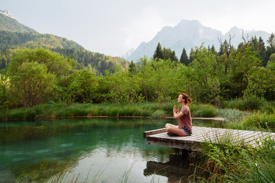 A woman practices outdoor meditation on a wooden boardwalk beside an emerald alpine lake. Harmonious eco-tourism concept focusing on spiritual connection with beautiful natural landscapes.