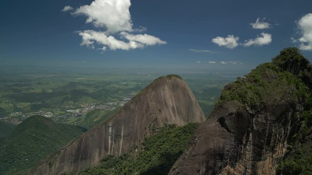 Panoramic aerial glide passing over Dedo de Deus mountain peaks, revealing green hills and Teres&oacute;polis city extending towards the horizon on deep blue sky day. Shot at 4K at 25 fps.