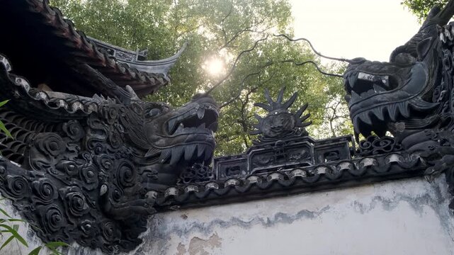 Sunlight filters through tree branches above two ornate carved dragon heads facing each other atop a traditional Chinese garden wall.