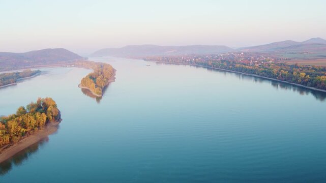Aerial drone footage flying over the wide Danube river, passing a long forested island towards a traditional village surrounded by hills at dusk.