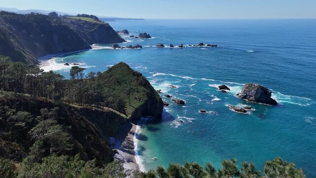 Aerial view elevating over pine trees revealing cliffs and turquoise sea of Asturian coastline in Spain