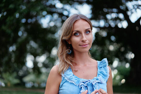 A relaxed outdoor portrait of a young woman wearing a ruffled blue dress, looking calmly at the camera against a soft leafy bokeh background, conveying natural beauty, serenity and summer vibes.