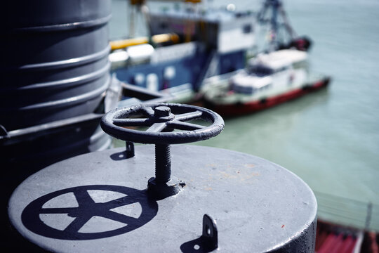 Close-up of metal ship valve on a deck hatch casting a strong shadow, with a blurred tugboat and harbor water in the background, evoking marine industry, nautical mechanics and industrial atmosphere.