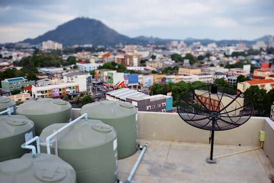 Rooftop view of Phuket city with water tanks, satellite dish, houses and mountain backdrop in soft tilt-shift focus.