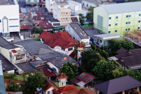 Aerial tilt-shift photograph of dense residential neighborhood featuring red tiled roofs, mixed houses and abundant trees, creating miniature effect that emphasizes urban architecture