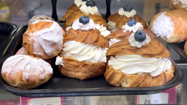 Freshly baked cream puff pastries generously filled with whipped cream, decorated with fresh blueberries, and arranged in a bakery display. Ideal for themes of desserts, pastry shops.