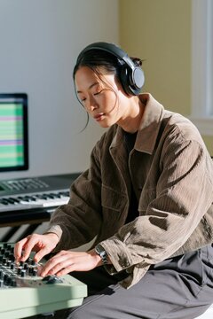 Female music producer adjusting mixer with headphones in home studio with keyboard and computer for audio production and beatmaking background