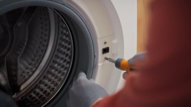 A specialist in work uniform disassembles the body of a front-loading washing machine unscrewing bolts in a bathroom next to a toolbox. Concept of technical maintenance and appliance diagnostics