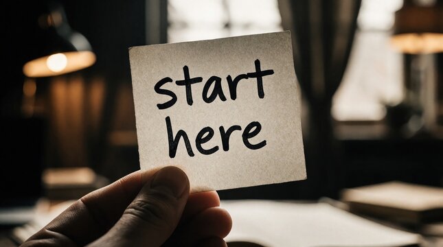 Hand holds paper note with start here text. Blurred desk background implies new beginnings, personal growth, and motivation. A clear direction for a journey.
