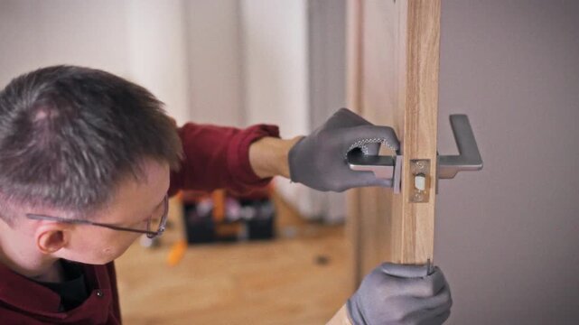 A close-up of gloved hands and a face in profile of a specialist repairing a door handle with an orange screwdriver on a wooden interior door. Concept of home repair and door lock maintenance