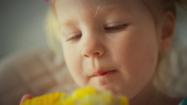 A child girl bites a ripe boiled corn cob while holding a kachan, captured in a close-up that shows the harvest-season setting inside a home kitchen, with basic food preparation process