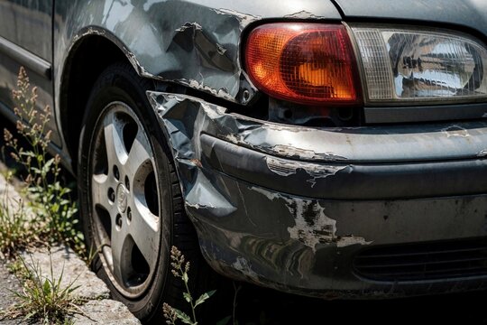 Damaged gray car rear bumper and headlight after crash. Metal body shows deep dents and peeling paint. Vehicle needs auto repair after accident on street.