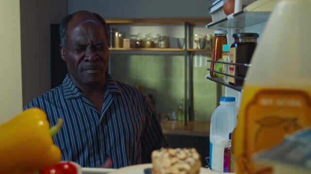 Senior African American man opening refrigerator, yawning, and smiling while taking plate of cake, POV. Ideal for ads about late-night cravings, unhealthy eating habits, or senior lifestyle