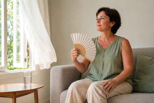 Mature woman managing menopause hot flash with hand fan at home